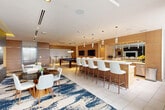 Located at 101 E Camino Real in Boca Raton, FL, this generously sized amenity lounge offers a refined, contemporary setting designed for socializing, entertaining, and productive gatherings. The open-plan layout is anchored by striking navy and cream abstract-patterned carpets that define distinct functional zones while lending the space a cohesive, upscale aesthetic.

The main lounge area features plush seating arrangements, including a deep-blue sectional sofa and curved accent chairs, centered around a wall-mounted TV set against warm wood-panel cladding. A bold fireplace feature wall adds visual drama. Natural light pours in through floor-to-ceiling windows, with views to the surrounding Boca Raton streetscape and access to an adjacent outdoor terrace.

The entertainment zone houses a full billiards table and high-top bar seating, while a large, well-appointed catering kitchen provides an expansive white-quartz island with bar stools, warm wood cabinetry, stainless steel appliances, and elegant pendant lighting — ideal for hosting events.

A glass-enclosed private meeting or dining room offers an intimate setting with a conference table, lounge seating, a wall-mounted display, and curated artwork. Throughout, tile flooring, recessed lighting, and mirrored surfaces reinforce the space's polished, modern character.