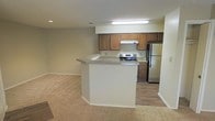 This single-floor residence offers a practical and comfortable open-concept layout that seamlessly connects the kitchen and living areas. The kitchen is a functional centerpiece, outfitted with warm honey-toned wood cabinetry that provides generous storage space, a stainless steel refrigerator, and a stainless steel range with an overhead ventilation hood. Light-toned countertops complement the cabinetry, while durable wood-look flooring grounds the cooking space with a clean, modern feel. A wraparound peninsula with a breakfast bar serves as a natural divider between the kitchen and the adjacent living area, offering additional prep space and casual seating. The living area transitions smoothly from the kitchen, featuring plush wall-to-wall carpeting that adds warmth and comfort underfoot. Neutral wall tones throughout create a bright, cohesive atmosphere that is easy to personalize. Recessed lighting in the living area and a fluorescent overhead fixture in the kitchen ensure the space is well-lit. The open flow between rooms enhances the sense of spaciousness, making this single-level layout both efficient and inviting for everyday living.