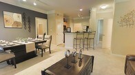 This single-floor residence showcases an open-concept design that integrates the living, dining, and kitchen areas into one cohesive, flowing space. The living area features plush carpeting in a warm neutral tone, complemented by recessed lighting and crown molding that add a refined architectural detail to the interior. A dark-finished coffee table serves as a focal point in the living space, accented by decorative metallic objects that contribute a contemporary aesthetic.

The dining area is positioned adjacent to the living room, furnished with elegant upholstered chairs and a sleek dining table, set against a charcoal accent wall that provides a striking contrast to the otherwise soft, neutral color palette throughout the space.

The kitchen is elevated by a breakfast bar with stylish wrought-iron barstools, offering both functional seating and a visual transition between the kitchen and living areas. Track lighting illuminates the kitchen workspace, which includes cabinetry in a warm wood finish and dark countertops. A hallway visible in the background suggests additional rooms beyond the main living area, extending the overall functionality of this well-appointed, single-level home.
