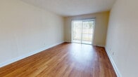 This single-floor residence offers a practical and comfortable layout with consistent warm-toned hardwood-style flooring throughout the main living areas. The open living room is generously proportioned, featuring neutral cream-colored walls, a textured ceiling, and a large sliding glass door fitted with vertical blinds that floods the space with natural light and provides direct access to the outdoors. Adjacent to the living area, a secondary open space flows seamlessly toward the kitchen, which is outfitted with white cabinetry, dark countertops, a double sink, and white appliances including a refrigerator, electric range, and dishwasher — all complemented by under-cabinet ventilation and ample storage. The bedroom is carpeted for comfort and includes a dedicated closet with built-in shelving, offering practical storage solutions. The bathroom is clean and functional, featuring a tiled shower-tub combination with an overhead shower, a window for natural ventilation, and wood-style flooring that ties the space together. Throughout the residence, the neutral color palette, consistent flooring, and efficient use of space create a cohesive and inviting atmosphere well-suited for comfortable everyday living.
