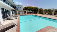 This outdoor amenity space is centered around a generously sized rectangular swimming pool with vivid turquoise water and brick-tiled coping, providing a clean, inviting aesthetic. The pool is enclosed by a black iron security fence, ensuring both safety and an open visual connection to the surrounding landscape. Stainless steel entry rails offer convenient pool access, while multiple lounge chairs positioned along the pool deck provide ample space for sunbathing and relaxation.

The multi-unit building forms a U-shaped backdrop around the pool area, with two levels of units featuring individual entry doors, private outdoor spaces, and exterior staircases connecting the floors. This layout creates a courtyard-like atmosphere that enhances the communal feel of the space.

Adjacent to the pool is a dedicated patio area shaded by large teal market umbrellas, furnished with round glass-top tables and sling-back chairs — ideal for outdoor dining or casual gatherings. BBQ grills are conveniently located nearby, complementing the outdoor entertaining setup. Mature trees line the perimeter, providing natural shade and greenery that softens the hardscape and adds to the overall pleasant, resort-like ambiance of the outdoor space.