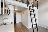 Located in Seattle's Roosevelt neighborhood, this two-level loft unit makes efficient use of its vertical space through a thoughtful, modern design. The main floor features an open-plan layout that seamlessly integrates the living, dining, and kitchen areas beneath soaring double-height ceilings. The kitchenette is neatly appointed with white cabinetry, a stainless steel compact refrigerator, microwave, and a single-basin sink — all designed for practical, streamlined living. Warm wood-look flooring runs throughout the main level, complementing the clean white walls and contemporary finishes.

A striking black metal ladder anchors the space visually and provides access to the upper loft level, which is outfitted with carpet and framed by a sleek black metal railing. The loft benefits from a row of horizontal windows that usher in ample natural light and a ceiling fan for added comfort. Large sliding glass doors on the main floor open the space further, inviting in natural light and views of the surrounding greenery.

The bathroom is efficiently designed with a dark-cabinet vanity, a wall-mounted mirror, and neutral flooring, maintaining the unit's consistent modern aesthetic. The entrance provides a clean transition into the main living area. Overall, this unit offers a clever, vertically-oriented layout well-suited for urban living in Seattle.