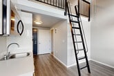 Located in Seattle's Roosevelt neighborhood, this two-level loft unit makes efficient use of its vertical space through a thoughtful, modern design. The main floor features an open-plan layout that seamlessly integrates the living, dining, and kitchen areas beneath soaring double-height ceilings. The kitchenette is neatly appointed with white cabinetry, a stainless steel compact refrigerator, microwave, and a single-basin sink — all designed for practical, streamlined living. Warm wood-look flooring runs throughout the main level, complementing the clean white walls and contemporary finishes.

A striking black metal ladder anchors the space visually and provides access to the upper loft level, which is outfitted with carpet and framed by a sleek black metal railing. The loft benefits from a row of horizontal windows that usher in ample natural light and a ceiling fan for added comfort. Large sliding glass doors on the main floor open the space further, inviting in natural light and views of the surrounding greenery.

The bathroom is efficiently designed with a dark-cabinet vanity, a wall-mounted mirror, and neutral flooring, maintaining the unit's consistent modern aesthetic. The entrance provides a clean transition into the main living area. Overall, this unit offers a clever, vertically-oriented layout well-suited for urban living in Seattle.