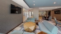This thoughtfully designed single-floor communal lounge offers a versatile, open layout that accommodates multiple seating configurations and activity zones. The space is defined by a sophisticated color palette of teal, gray, and warm wood tones, carried throughout the patterned carpet tile flooring and coordinated upholstered furnishings.

The main lounge area features a generous arrangement of teal sectional sofas and geometric-patterned accent chairs grouped around sculptural faceted wood coffee tables, creating an inviting setting for casual socializing. A wall-mounted flat-screen television anchors one side of the lounge, backed by textured gray wall covering. Floor-to-ceiling curtain-wall glazing along one side of the space floods the interior with natural light and provides views of an adjacent outdoor area.

A dedicated dining zone includes a round table with patterned side chairs alongside a tall communal bar-height counter with pendant lighting and barstool seating. Glass-partitioned rooms visible from the main area offer private spaces for meetings or focused work.

The kitchenette is finished with quartz countertops, a stainless steel undermount sink, gray cabinetry, a full-size stainless refrigerator, and a distinctive hexagonal tile backsplash, providing functional refreshment amenities. Recessed lighting throughout ensures a well-lit, welcoming atmosphere.