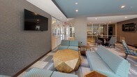 This thoughtfully designed single-floor communal lounge offers a versatile, open layout that accommodates multiple seating configurations and activity zones. The space is defined by a sophisticated color palette of teal, gray, and warm wood tones, carried throughout the patterned carpet tile flooring and coordinated upholstered furnishings.

The main lounge area features a generous arrangement of teal sectional sofas and geometric-patterned accent chairs grouped around sculptural faceted wood coffee tables, creating an inviting setting for casual socializing. A wall-mounted flat-screen television anchors one side of the lounge, backed by textured gray wall covering. Floor-to-ceiling curtain-wall glazing along one side of the space floods the interior with natural light and provides views of an adjacent outdoor area.

A dedicated dining zone includes a round table with patterned side chairs alongside a tall communal bar-height counter with pendant lighting and barstool seating. Glass-partitioned rooms visible from the main area offer private spaces for meetings or focused work.

The kitchenette is finished with quartz countertops, a stainless steel undermount sink, gray cabinetry, a full-size stainless refrigerator, and a distinctive hexagonal tile backsplash, providing functional refreshment amenities. Recessed lighting throughout ensures a well-lit, welcoming atmosphere.