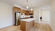 This single-floor residence offers a thoughtfully designed layout with clean, contemporary finishes throughout. The kitchen serves as the heart of the space, showcasing warm wood cabinetry, stainless steel appliances including a refrigerator, electric range, microwave, and dishwasher, all complemented by crisp white countertops. A central island with a matching white surface provides additional prep space and casual seating potential, while two pendant lights overhead add a stylish focal point. Light hardwood-style flooring flows seamlessly throughout the entire unit, creating a cohesive and airy feel. The living area is generously proportioned, bathed in natural light from large windows fitted with horizontal blinds, and features a door providing direct access to an outdoor space with tree-lined views. A dedicated in-unit laundry area is conveniently accessible from the kitchen corridor. The bedroom includes a spacious sliding-door closet with built-in shelving for ample storage. The bathroom is well-equipped with a full soaking tub and shower combination, brushed nickel fixtures, a wood-toned vanity with a white countertop, and matching hardwood-style flooring, maintaining the unit's consistent warm aesthetic throughout.