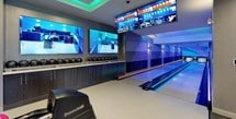 This dynamic entertainment space is designed to deliver an immersive recreational experience across several distinct areas. At the heart of the venue are professional-grade Brunswick bowling lanes, illuminated by vivid blue LED lighting that runs along the lane edges, with large mounted flat-screen displays overhead for live scoring and gameplay tracking. Dark cabinetry along the service area provides ample storage for a full selection of colorful bowling balls.

Adjacent to the lanes is a generously sized lounge area, finished with plush gray sectional sofas accented by bold blue and black decorative pillows. A tray ceiling fitted with teal LED cove lighting gives the space a contemporary, upscale ambiance, while wall-mounted sconces add warm, layered illumination. A mirrored mosaic coffee table serves as a striking centerpiece, and an eye-catching sculptural wall installation composed of metallic orbs adds a sophisticated artistic touch.

The connecting corridor and game room area features an air hockey table, bold pop-art wall décor with arcade-style graphics, and smooth transitions between spaces, all finished with neutral carpeting and recessed lighting. The overall design balances entertainment functionality with a polished, modern aesthetic throughout.