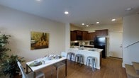 This two-story residence offers a thoughtfully designed layout that blends functionality with modern style. The open-concept kitchen and dining area serve as the heart of the home, creating a seamless flow between cooking, dining, and entertaining spaces. The kitchen features rich, dark wood cabinetry paired with a crisp white countertop peninsula, which doubles as a breakfast bar with industrial-style metal stools. Black appliances, including a full-size refrigerator and built-in microwave, add a sleek, contemporary touch. Recessed lighting illuminates the space evenly, enhancing the clean, bright atmosphere. The adjacent dining area accommodates a rectangular white dining table with modern seating, complemented by warm hardwood flooring that runs throughout the main level, adding depth and warmth to the overall aesthetic. A piece of contemporary wall art adds a subtle artistic accent to the neutral, warm-toned walls. The open layout allows natural sight lines across both the kitchen and dining zones, making the space feel expansive and welcoming. Spread across two floors, the residence offers ample room for comfortable everyday living, with a versatile and well-appointed design that suits a variety of lifestyles.