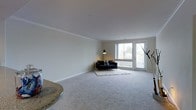 This well-appointed single-floor space offers a clean, modern aesthetic defined by neutral tones and an open, airy layout. The living area is generously proportioned, featuring soft wall-to-wall carpeting that adds warmth and comfort underfoot. Smooth white ceilings with elegant crown molding lend a polished, refined quality to the interior.

Natural light flows abundantly into the living area through a large picture window paired with full-length French doors, which provide direct access to an outdoor space and offer pleasant views of the surrounding greenery. The combination of these openings creates a bright, welcoming atmosphere throughout the day.

The living space is well-suited for a variety of furniture arrangements, with ample room to define distinct seating and lounging zones. A partial view of a granite-topped surface near the entrance suggests a seamlessly connected dining or kitchen area, hinting at an open-concept flow between the main living spaces.

Overall, the space presents a light-filled, move-in-ready environment with a calm and neutral palette, making it highly versatile and easy to personalize. The thoughtful balance of natural light, durable finishes, and open space creates an inviting and functional everyday living environment.