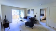 This single-floor space features an open-concept living area bathed in natural light, courtesy of large sliding glass doors that lead to a private outdoor balcony. The balcony extends the living space outdoors and offers views of surrounding greenery, making it a notable feature of the unit. The living room is carpeted and furnished with a plush blue sofa and a coordinating accent armchair with nailhead detailing, arranged around a sleek hairpin-leg coffee table. Two framed art pieces adorn the neutral-toned walls, adding a decorative touch. A side table with a lamp provides ambient lighting. Adjacent to the living room, an open doorway connects directly to the bedroom, which features a large bed with an upholstered headboard and continued carpeted flooring, maintaining a cohesive aesthetic throughout. The neutral wall palette and light-colored carpet create a clean, airy atmosphere. The layout emphasizes ease of flow between spaces, with the living area serving as the central hub connecting both the outdoor balcony and the sleeping quarters.