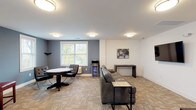 The amenity lounge at Meridia Transit Crossing, Building B, is a well-appointed, single-floor communal space designed to foster social interaction and relaxation among residents. The room features a neutral color palette with crisp white walls accented by slate-blue feature walls, creating a modern yet inviting atmosphere throughout.

Patterned carpet tile flooring runs throughout the space, adding texture and warmth underfoot while providing durability for high-traffic use. Flush-mount ceiling light fixtures provide consistent, even illumination across the entire area.

The lounge is divided into distinct functional zones. A dedicated game area occupies one corner, centered around a round game table with cushioned chairs, making it well-suited for card games or casual gatherings. Adjacent to the game table, a compact beverage cooler offers added convenience for entertaining.

The main seating area features a large leather sectional sofa oriented toward a wall-mounted flat-screen television, creating a comfortable media-viewing space. Sleek console tables and side tables complement the furnishings throughout.

Additional seating is provided in a secondary alcove with red upholstered bar-height chairs and a decorative mirror, adding a refined touch. Multiple windows allow natural light to filter into the space, offering views of the surrounding outdoor environment and enhancing the overall ambiance of the lounge.