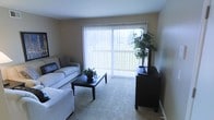 This single-story space features a welcoming living room designed for comfort and functionality. The room is anchored by a large sliding glass door dressed with vertical blinds, allowing abundant natural light to fill the space while offering views of the outdoors, including surrounding greenery. The neutral-toned walls and plush carpeting contribute to a calm, understated ambiance throughout.

The living room is thoughtfully arranged with a light-colored sofa and a matching chair, positioned around a dark-finished rectangular coffee table that serves as the room's focal point. A coordinating dark wood media console sits adjacent to the sliding door, topped with a flat-screen television and a decorative potted plant that adds a touch of natural warmth. Additional greenery placed on the coffee table further enhances the inviting atmosphere.

The overall design aesthetic is clean and practical, with a neutral color palette of beige and warm gray tones that lend a cohesive, livable feel. The space balances everyday functionality with comfortable living, making it well-suited for relaxed daily use.