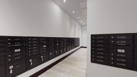 This mailroom serves as a centralized mail distribution hub within a multi-unit building. The space is designed with functionality and clarity in mind, featuring an extensive array of matte black individual mailboxes mounted along the walls, each clearly labeled with unit identifiers spanning multiple building sections — including E and W unit designations across numerous floors. The mailboxes are arranged in orderly rows, making navigation straightforward for residents.

The room's interior design is clean and modern, characterized by crisp white walls that contrast sharply with the dark mailbox units. The light hardwood-style flooring adds a refined touch to the utilitarian space, while a dark baseboard trim provides a grounded visual boundary along the wall line. Recessed ceiling lights are evenly distributed throughout, ensuring the room is well-lit and easy to use at any time of day.

The layout is corridor-style, with mailboxes lining at least two wall surfaces, maximizing the number of units that can be accommodated. A secondary panel of smaller mailboxes is visible on an adjacent wall, further expanding capacity. The overall ambiance is tidy, professional, and efficiently organized, reflecting thoughtful design suited to a high-occupancy residential or mixed-use building.