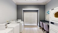 This community laundry facility in Columbia, SC, is a clean and functional space designed to serve residents efficiently. The room is finished with light tile flooring and two-tone walls in soft gray and slate, accented by crisp white crown molding that lends a polished look throughout. Overhead, a flush panel light fixture provides bright, even illumination across the entire space.

The facility is equipped with multiple top-load washing machines and stacked dryer units, offering ample capacity for simultaneous use. A dedicated folding and seating area features a dark countertop surface accompanied by two upholstered lavender chairs, providing a comfortable spot to wait. A community bulletin board mounted above the counter displays washing and drying instructions, keeping residents informed and the space organized.

A large, deeply recessed window fitted with horizontal blinds allows natural light to filter in while maintaining privacy. The room is accessed through a classic white French door with glass panels, adding a touch of elegance to the utilitarian setting. Safety is addressed with a prominently mounted fire extinguisher near the entrance. The space flows logically, with machines arranged along the walls to maximize open floor space and ease of movement, making this laundry room both practical and comfortable for everyday use.