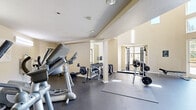 Located at 3560 Flora Vista Ave in Santa Clara, CA, this fitness facility offers a thoughtfully designed exercise space that combines functionality with an open, airy atmosphere. The exercise room is distinguished by soaring ceilings with a clerestory band of windows that flood the interior with natural light, creating a bright and energizing environment. The durable rubber flooring provides a safe, supportive surface throughout the workout area. Equipment includes treadmills, elliptical trainers, a cable weight machine, a free weight bench, and a full dumbbell rack, catering to both cardio and strength training needs. Wall-mounted mirrors expand the sense of space, while a mounted flat-screen TV adds convenience for longer sessions. A staircase leads to an upper-level mezzanine area, adding vertical dimension to the layout. Large glass doors open directly onto an expansive outdoor terrace paved with decorative tile. The terrace features a raised wood platform deck, potted palms, and shaded seating areas beneath large umbrellas, all set against a backdrop of mature trees and clear California skies. An outdoor water station and additional patio space complete this well-rounded fitness amenity, ideal for an active lifestyle in the heart of Santa Clara.