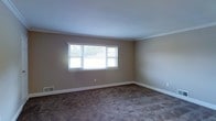 This single-floor home presents a clean, straightforward layout well-suited for comfortable everyday living. The interior is finished with neutral beige walls that create a warm, understated ambiance throughout the space. Crown molding lines the ceilings, adding a classic architectural detail that lends a sense of polish to the rooms. The flooring is covered in wall-to-wall carpet in a muted, earthy tone, providing a soft and quiet underfoot experience. Natural light enters through multi-pane windows dressed with horizontal blinds, brightening the interior while allowing for privacy control. The windows offer views of the surrounding outdoor greenery, connecting the interior to the natural environment outside. White baseboards run along the perimeter of the rooms, complementing the neutral color palette and providing a crisp contrast against the carpeted floors. Ventilation is provided through floor-level registers, indicating a central HVAC system for year-round climate control. The overall atmosphere is calm and move-in ready, with a modest and functional design that provides a solid foundation for personalization and furnishing.