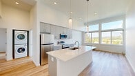 Located at 7460 SW Hermoso Way in Tigard, Oregon, this thoughtfully designed 2-bedroom, 2-bathroom residence offers a contemporary living experience on a single floor. The open-concept layout is anchored by a striking kitchen featuring a large white quartz island with an undermount sink, sleek gray cabinetry, stainless steel appliances, and elegant pendant lighting hanging from soaring high ceilings. The kitchen flows seamlessly into the living area, which is bathed in natural light from an expansive wall of floor-to-ceiling windows and a glass door opening to a private balcony with scenic views of lush, mature trees and the surrounding Tigard landscape. Light hardwood flooring runs throughout the entire residence, enhancing the bright and airy atmosphere. The spacious primary bedroom offers a generous closet and direct access to a hallway connecting the rest of the unit. Both bathrooms are finished in a clean, modern style with gray vanity cabinetry, chrome fixtures, tiled surround tub-shower combinations, and wood-look flooring. The secondary bedroom is intimate yet comfortable, featuring a window with a peaceful wooded view. The hallway connects all rooms efficiently, completing this well-planned, contemporary home.