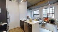 Studio 606 at 121 Westmoreland Avenue in White Plains, NY, is a thoughtfully designed loft-style studio that blends industrial character with modern finishes. The space is defined by its soaring exposed concrete ceilings with raw wooden beams and visible ductwork, lending an authentic warehouse aesthetic throughout. Large, multi-pane industrial-style windows flood the open-concept living area with abundant natural light while offering sweeping views of the White Plains skyline.

The kitchen is fully equipped with stainless steel appliances — including a refrigerator, electric range, and built-in microwave — complemented by two-tone cabinetry in white and dark wood, white quartz countertops, and a peninsula with bar seating for two, ideal for casual dining. The open layout seamlessly connects the kitchen to the combined living and sleeping area, where wood-plank flooring runs throughout, enhancing warmth and continuity.

The living and sleeping zone is generously proportioned, accommodating both a full bedroom setup and a separate lounge area with ample room to personalize. A dedicated bathroom features neutral tile flooring, soft grey walls, and clean, modern fixtures. The overall ambiance is urban and sophisticated, balancing the raw, industrial bones of the building with contemporary design elements for a stylish and functional living experience.