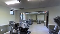 This single-floor fitness center offers a thoughtfully designed workout space that balances functionality with a clean, modern aesthetic. The room features light hardwood-style flooring throughout, complemented by soft green-gray accent walls and white trim that create a calm, focused atmosphere. Large windows along one wall invite natural light and offer views of surrounding greenery, enhancing the overall ambiance of the space. Ceiling fans are mounted overhead to maintain comfortable airflow during workouts. A full-length mirrored wall visually expands the room and provides essential feedback for exercise form, while also reflecting the natural light and lending the space a more open feel. The fitness center is equipped with a variety of cardio machines, including stationary bikes and a stair climber, as well as multiple strength training stations positioned throughout the room. Storage shelving is conveniently placed within the space. Safety is addressed with a clearly visible fire extinguisher and an illuminated emergency exit sign. The combination of practical equipment, thoughtful layout, and a welcoming interior design makes this fitness center a well-rounded facility suitable for a range of workout routines.