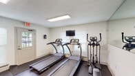 This fitness center in Baton Rouge, Louisiana, offers a comprehensive workout environment designed to accommodate a variety of exercise routines. The space is organized into distinct functional zones, making efficient use of its single-floor layout. Flooring throughout is dark rubber, providing durability and traction suited to an active environment. Recessed rectangular LED flush-mount panels illuminate the space evenly, and a textured ceiling adds a clean, finished look overhead.

The cardio zone features two SportsArt treadmills and an elliptical trainer positioned along one wall, with windows fitted with white blinds allowing natural light to filter in. A wall-mounted flat-screen television is centrally placed for viewing during cardio sessions. This area also includes a stationary recumbent bicycle, offering low-impact exercise options.

The strength training area is anchored by a multi-functional cable machine, providing a wide range of resistance exercises. A rack of dumbbells lines the wall alongside a weight bench, catering to free weight workouts. A stability ball is also available for core training. A full-length wall mirror spans one side of the strength zone, enhancing the sense of openness while allowing users to monitor their form. The fitness center is accessible via a keyed entry door, ensuring a secure and dedicated workout space.