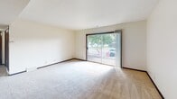 This single-floor residential unit offers a functional and straightforward layout designed for comfortable everyday living. The living area is generously sized, featuring wall-to-wall carpeting, neutral white walls, and a large sliding glass door that opens to a private balcony, allowing abundant natural light and outdoor views of surrounding greenery. A distinctive built-in open shelving unit serves as a subtle architectural divider within the living space, adding both character and practical storage.

The kitchen is efficiently laid out with warm-toned wood cabinetry featuring decorative scalloped trim, a double sink, white appliances including a refrigerator and dishwasher, and durable hardwood-style flooring. The transition from the kitchen to the carpeted living areas is clean and well-defined.

The bedroom is a comfortable, carpeted room with a large window fitted with vertical blinds, offering natural light while maintaining privacy. Dark wood door frames and trim are consistent throughout the unit, providing a cohesive design element.

The bathroom is clean and practical, featuring a vanity with a granite-style countertop, a toilet, and a full bathtub with shower. Light tile flooring completes the space. Overall, the unit presents a clean, move-in-ready environment with a neutral palette throughout.