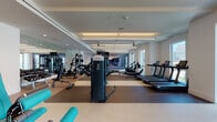 This contemporary fitness center is designed to offer a complete workout experience within a thoughtfully organized, spacious layout. The facility is divided into distinct training zones, each purpose-built to accommodate different exercise disciplines. The cardio area features a row of treadmills, ellipticals, and stationary bikes, all positioned alongside large floor-to-ceiling windows that invite abundant natural light and offer impressive views of the surrounding cityscape. A set of glass doors opens to an outdoor balcony, further enhancing the connection between the interior workout space and the urban environment beyond. The strength training zone is outfitted with a broad selection of cable machines and selectorized weight equipment, finished in a striking teal-and-black color scheme. Full-length mirrors line the walls throughout the facility, visually expanding the space and allowing users to monitor their form. The flooring transitions between rubber and smooth surfaces to suit different training activities. Overhead, linear LED lighting is recessed into coffered ceilings, complemented by ceiling fans that maintain air circulation. The overall ambiance is clean, bright, and professional, reflecting a high standard of fitness amenity design.