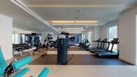 This contemporary fitness center is designed to offer a complete workout experience within a thoughtfully organized, spacious layout. The facility is divided into distinct training zones, each purpose-built to accommodate different exercise disciplines. The cardio area features a row of treadmills, ellipticals, and stationary bikes, all positioned alongside large floor-to-ceiling windows that invite abundant natural light and offer impressive views of the surrounding cityscape. A set of glass doors opens to an outdoor balcony, further enhancing the connection between the interior workout space and the urban environment beyond. The strength training zone is outfitted with a broad selection of cable machines and selectorized weight equipment, finished in a striking teal-and-black color scheme. Full-length mirrors line the walls throughout the facility, visually expanding the space and allowing users to monitor their form. The flooring transitions between rubber and smooth surfaces to suit different training activities. Overhead, linear LED lighting is recessed into coffered ceilings, complemented by ceiling fans that maintain air circulation. The overall ambiance is clean, bright, and professional, reflecting a high standard of fitness amenity design.