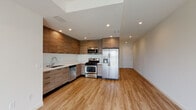 This contemporary single-floor unit is designed with a clean, modern aesthetic that carries consistently throughout every room. The open-concept kitchen serves as the heart of the space, showcasing floor-to-ceiling wood-grain cabinetry, a crisp white countertop with a undermount sink, and a full suite of stainless steel appliances including a gas range, built-in microwave, dishwasher, and a French-door refrigerator. Recessed lighting illuminates the kitchen and living areas, complementing the warm, light-toned hardwood-style flooring that extends seamlessly across the entire unit. The living area is bright and open, offering a generous, flexible layout. The bedroom features a mirrored sliding-door closet providing ample storage, with the same warm flooring continuing throughout. The unit includes two bathrooms: one featuring a tub-shower combination with sliding glass doors and wood-tone vanity, and a second with a standalone glass-enclosed shower stall and an extended double vanity topped with a wide mirror. Clean white walls, recessed lighting, and cohesive wood-tone finishes across cabinetry and vanities create a unified, contemporary atmosphere throughout the entire space.
