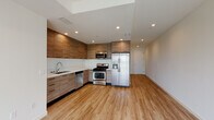 This contemporary single-floor unit is designed with a clean, modern aesthetic that carries consistently throughout every room. The open-concept kitchen serves as the heart of the space, showcasing floor-to-ceiling wood-grain cabinetry, a crisp white countertop with a undermount sink, and a full suite of stainless steel appliances including a gas range, built-in microwave, dishwasher, and a French-door refrigerator. Recessed lighting illuminates the kitchen and living areas, complementing the warm, light-toned hardwood-style flooring that extends seamlessly across the entire unit. The living area is bright and open, offering a generous, flexible layout. The bedroom features a mirrored sliding-door closet providing ample storage, with the same warm flooring continuing throughout. The unit includes two bathrooms: one featuring a tub-shower combination with sliding glass doors and wood-tone vanity, and a second with a standalone glass-enclosed shower stall and an extended double vanity topped with a wide mirror. Clean white walls, recessed lighting, and cohesive wood-tone finishes across cabinetry and vanities create a unified, contemporary atmosphere throughout the entire space.