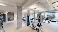 This well-appointed fitness center offers a generously sized, single-level layout designed to accommodate a full spectrum of workout needs. The space features a contemporary design with neutral-toned carpeted flooring, coffered ceilings with recessed and linear pendant lighting, and structural columns finished in a warm stone-like veneer that lend an upscale aesthetic throughout.

The main exercise floor is densely equipped with a wide variety of professional-grade strength machines, free weights, benches, and cable systems, providing options for both resistance and functional training. A dedicated rack of dumbbells is neatly organized along one wall, while additional equipment such as rowing machines and cardio stations are positioned to maximize floor space and traffic flow.

Along the perimeter, large windows flood the space with natural light and offer views of the surrounding exterior, creating an open and energizing atmosphere. A separate studio area features full-length mirrors, open floor space, and TRX suspension training equipment, making it ideal for yoga, stretching, or group fitness activities.

Additional amenities include a water fountain station flanked by decorative potted plants, and a glass-enclosed room that adds a refined architectural element. The overall ambiance is clean, modern, and professional, reflecting a high standard of facility design suited for serious fitness use.