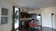 This single-floor space showcases a well-appointed kitchen and adjacent dining area designed with a contemporary aesthetic. The kitchen is anchored by rich, dark-stained cabinetry that extends to the upper walls, providing ample storage space. Granite countertops in complementary dark tones run throughout, offering generous preparation surfaces. Stainless steel appliances, including a refrigerator, electric range with microwave above, and dishwasher, lend a polished, cohesive look to the space.

A built-in breakfast bar extends from the kitchen, furnished with sleek bar-height seating featuring bold red upholstered chairs, creating a striking contrast against the darker cabinetry and flooring. Track lighting runs along the ceiling, providing targeted illumination throughout the kitchen and dining area.

Dark hardwood flooring flows throughout the space, adding warmth and continuity. A large window framed with dark curtain panels allows natural light to fill the area, complemented by a tall indoor plant that introduces a natural element. The overall ambiance balances modern functionality with warm, inviting design details.