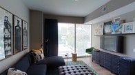 This single-floor space showcases a thoughtfully designed living area that balances modern aesthetics with eclectic character. The room is anchored by expansive floor-to-ceiling windows fitted with horizontal blinds, flooding the interior with natural light while framing views of surrounding urban greenery and cityscape beyond. Dark floor-length curtains flank the windows, adding a dramatic contrast to the warm, taupe-toned walls.

The living area features light hardwood-style flooring that complements the neutral palette throughout. A deep, dark sectional sofa adorned with patterned throw pillows serves as the central seating element, paired with a tufted charcoal ottoman that doubles as a coffee table surface. A petite round side table and a natural wood stump accent piece add organic touches to the arrangement.

A vibrant emerald green accent chair introduces a bold pop of color near the window, contributing to the room's eclectic personality. Along one wall, a dark navy dresser provides storage and display space alongside a mounted flat-screen television. A geometric-patterned area rug in teal and yellow grounds the seating arrangement, tying together the room's mix of colors and textures into a cohesive and visually engaging living environment.