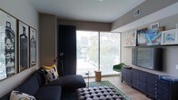 This single-floor space showcases a thoughtfully designed living area that balances modern aesthetics with eclectic character. The room is anchored by expansive floor-to-ceiling windows fitted with horizontal blinds, flooding the interior with natural light while framing views of surrounding urban greenery and cityscape beyond. Dark floor-length curtains flank the windows, adding a dramatic contrast to the warm, taupe-toned walls.

The living area features light hardwood-style flooring that complements the neutral palette throughout. A deep, dark sectional sofa adorned with patterned throw pillows serves as the central seating element, paired with a tufted charcoal ottoman that doubles as a coffee table surface. A petite round side table and a natural wood stump accent piece add organic touches to the arrangement.

A vibrant emerald green accent chair introduces a bold pop of color near the window, contributing to the room's eclectic personality. Along one wall, a dark navy dresser provides storage and display space alongside a mounted flat-screen television. A geometric-patterned area rug in teal and yellow grounds the seating arrangement, tying together the room's mix of colors and textures into a cohesive and visually engaging living environment.