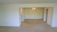 This single-floor space features an open, flowing layout characterized by clean white walls and neutral-toned wall-to-wall carpeting throughout. The main living area is generously sized, offering ample room for flexible furniture arrangements. A wide, open archway connects the primary living space to an adjoining area, enhancing the sense of openness and allowing for easy movement between zones. A ceiling fan with an integrated light fixture is centrally mounted, providing both air circulation and overhead lighting. A standard entry door is visible along the far wall, offering direct access to the space. A partial wall divider adds subtle architectural definition without fully enclosing the areas, maintaining the open feel. An adjacent area, partially visible through the archway, suggests additional functional space, potentially suitable for dining or secondary use. The overall ambiance is clean, neutral, and move-in ready, providing a blank canvas that can accommodate a variety of interior styles and configurations.