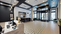 This single-floor lobby presents a sophisticated and well-appointed common area designed with both form and function in mind. The space immediately draws attention with its striking black-and-white geometric patterned tile flooring, which anchors the room and establishes a bold visual identity. Recessed lighting and modern pendant fixtures with matte black finishes illuminate the interior, complementing the dark structural beam accents that frame the ceiling and add architectural contrast against the white walls.

A prominent feature is the sleek, dark-toned bar or lounge counter fitted with black cabinetry and gold-accented bar stools, accompanied by an open display shelf stocked with decorative items and bottles. A decorative globe adds a classic, worldly touch to the reception counter area on the left side of the space.

Floor-to-ceiling glass entry doors on the right side of the lobby allow abundant natural light to pour in, creating an open and inviting atmosphere. A glimpse of a furnished lounge area with green upholstered seating is visible in the background, hinting at a comfortable social space just beyond the main entrance area. Overall, the lobby conveys a polished, contemporary ambiance.