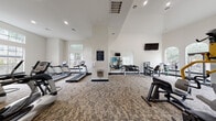 This fitness center is designed to support a comprehensive workout experience within a single-level, generously proportioned space. The facility is characterized by soaring vaulted and high ceilings that lend an open, airy feel throughout. Large arched windows line key areas of the room, flooding the interior with abundant natural light and offering views of the surrounding greenery outdoors.

The main workout area is divided into distinct zones for cardio and strength training. The cardio section is equipped with multiple treadmills, elliptical machines, and stationary bikes, arranged along windowed walls to create an inviting environment for endurance training. A dedicated strength training area features a full dumbbell rack with a range of free weights, several multi-function cable and resistance machines, and additional weight equipment to support varied training routines.

A large wall mirror expands the sense of space in the strength zone, while multiple wall-mounted flat-screen televisions are positioned throughout the facility for entertainment during workouts. A built-in water fountain provides convenient hydration access. The floors are covered in a patterned carpet that adds texture and comfort underfoot. Decorative blue abstract artwork adorns the walls, contributing a polished, modern aesthetic. The overall ambiance is clean, bright, and well-organized, making it a functional and welcoming environment for fitness.