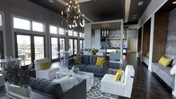 This sophisticated single-floor space makes an immediate impression with its dramatic design aesthetic. The main lounge area features soaring ceilings finished in a deep charcoal tone, anchored by a striking wrought-iron chandelier with candelabra-style lighting that serves as a true focal point. Large floor-to-ceiling windows and glass doors flood the space with abundant natural light while offering views to an exterior terrace, creating a seamless indoor-outdoor connection. The seating area is thoughtfully arranged with plush grey tufted sofas and crisp white accent chairs, all unified by a bold grey-and-white patterned area rug. Pops of chartreuse yellow in the throw pillows and accent chairs add a vibrant, modern energy throughout. Towards the rear, a well-appointed bar and lounge area is visible, featuring pendant lighting, open shelving, and a sleek counter — ideal for entertaining. Dark hardwood flooring runs throughout, complementing the overall moody yet refined color palette of greys, whites, and yellows. The open-plan layout encourages a fluid flow between the lounge and bar zones, making this a highly functional and visually cohesive space designed for both comfort and style.
