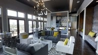 This sophisticated single-floor space makes an immediate impression with its dramatic design aesthetic. The main lounge area features soaring ceilings finished in a deep charcoal tone, anchored by a striking wrought-iron chandelier with candelabra-style lighting that serves as a true focal point. Large floor-to-ceiling windows and glass doors flood the space with abundant natural light while offering views to an exterior terrace, creating a seamless indoor-outdoor connection. The seating area is thoughtfully arranged with plush grey tufted sofas and crisp white accent chairs, all unified by a bold grey-and-white patterned area rug. Pops of chartreuse yellow in the throw pillows and accent chairs add a vibrant, modern energy throughout. Towards the rear, a well-appointed bar and lounge area is visible, featuring pendant lighting, open shelving, and a sleek counter — ideal for entertaining. Dark hardwood flooring runs throughout, complementing the overall moody yet refined color palette of greys, whites, and yellows. The open-plan layout encourages a fluid flow between the lounge and bar zones, making this a highly functional and visually cohesive space designed for both comfort and style.