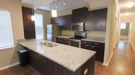 This well-designed single-floor space showcases a contemporary aesthetic with thoughtful finishes throughout. The kitchen is a standout feature, boasting rich dark espresso cabinetry that extends to the ceiling, offering generous storage. Light granite countertops with subtle veining provide a striking contrast against the dark cabinetry and are carried across both the perimeter counters and a large central island. The island, equipped with a double-basin stainless steel sink, serves as a functional centerpiece ideal for meal preparation and casual dining. Stainless steel appliances — including a refrigerator, range, and over-the-range microwave — lend a polished, modern look. Overhead, a combination of pendant lights and track lighting illuminates the kitchen with both warmth and practicality. Natural light filters into the space through a window fitted with blinds, enhancing the open and airy atmosphere. The warm hardwood-style flooring ties the space together, complementing the overall contemporary palette. The clean lines, quality materials, and cohesive design make this a highly functional and visually appealing space.