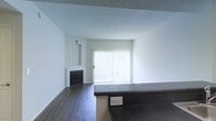 This single-floor space showcases a clean, open-concept layout designed for comfortable everyday living. The main living area is anchored by a classic fireplace with a framed surround, adding a focal point to the room and providing a cozy atmosphere. Dark hardwood flooring runs throughout, creating a rich contrast against the crisp white walls and adding a sense of warmth and sophistication to the interior. A large sliding glass door dominates the far wall, fitted with vertical blinds that allow for adjustable light control. When open, this feature floods the living space with abundant natural light and provides access to an outdoor area. The textured ceiling adds subtle architectural character to the room. A dark countertop surface is visible in the foreground, suggesting a kitchen or bar area that flows directly into the living space, reinforcing the open-plan feel. The neutral white walls offer a versatile backdrop suitable for a variety of décor styles. Overall, the space presents a bright, functional, and well-appointed interior with a balanced mix of practical features and clean design elements.