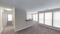 This single-floor space showcases a thoughtfully designed open layout that promotes a seamless flow between living areas. The interior is finished with soft, neutral-toned walls that create a clean and versatile backdrop throughout. Plush wall-to-wall carpeting runs across the main living area, adding warmth and comfort underfoot.

A defining architectural feature is the partial dividing wall topped with a dark-finished breakfast bar, which elegantly separates the kitchen area from the living space while maintaining an open, connected feel. A pendant light hangs above the bar, providing focused task lighting and a subtle design accent.

Crown molding frames the ceilings, lending a polished, classic character to the space. Large windows with vertical blinds and additional horizontal blinds allow for ample natural light to pour in, brightening the interior while offering flexible light control. The windows also provide a sense of openness and an airy ambiance throughout.

Overall, the space combines practical design elements with refined finishing touches, resulting in a bright, neutral, and move-in-ready environment well-suited for comfortable everyday living.
