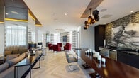 The Laureat Clubhouse in Fort Lauderdale delivers a refined, resort-inspired amenity experience across a single, expansive floor. The interior design blends contemporary elegance with warm, residential comfort, anchored throughout by light herringbone-patterned hardwood flooring that unifies each distinct zone seamlessly.

The lounge area serves as a social centerpiece, furnished with richly upholstered crimson armchairs arranged around a tufted ottoman beneath a striking sputnik-style chandelier. Floor-to-ceiling windows dressed in bold striped drapery flood the space with natural light and provide views to an adjacent outdoor terrace.

A dedicated entertainment zone features a sleek billiards table set atop a dramatic dark area rug, accompanied by a stylish wet bar with marble countertops, dark cabinetry, and sculptural pendant lighting. Nearby, a media lounge offers modular blue-gray sofas facing a built-in entertainment wall with illuminated shelving and a wall-mounted television beneath a rich dark-paneled ceiling.

A formal dining and conference area includes a long dark wood table with modern chrome-accented seating, ideal for meetings or private gatherings. Glass-partitioned walls define a quieter seating nook, while the open corridor layout ensures smooth flow between areas. The overall ambiance is polished, inviting, and thoroughly contemporary.