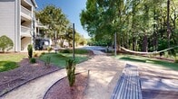 Located at 1000 Park Place Ave in Morrisville, NC, this thoughtfully designed outdoor amenity space offers residents a serene and well-appointed retreat surrounded by nature. The grounds are framed by a dense canopy of mature trees — including tall pines and leafy hardwoods — that provide ample shade and a tranquil, wooded ambiance throughout the space.

A winding pathway connects the various activity zones, guiding visitors through landscaped beds of mulch, trimmed shrubs, and patches of lush green turf. Suspended hammocks are strung between upright poles, offering comfortable spots to unwind beneath the tree canopy. Nearby, a cluster of Adirondack-style chairs is arranged around a central gathering point, ideal for socializing or quiet relaxation.

Wooden bench seating and thatched umbrella structures are strategically placed throughout, adding both function and aesthetic character. String lights are strung between poles, hinting at an inviting atmosphere during evening hours. The adjacent multi-story residential building features traditional architectural detailing, private balconies, and well-maintained landscaping that seamlessly integrates with the outdoor community space. Together, these elements create an engaging, resort-like environment that prioritizes comfort and community connection.