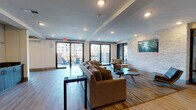 Located at 2421 Landmark Dr in Raleigh, NC, this single-floor amenity lounge is thoughtfully designed to serve as a stylish, multifunctional gathering space. The main lounge area is defined by wide-plank wood-look tile flooring, recessed lighting, and an eye-catching stacked stone accent wall that anchors a wall-mounted flat-screen television. Multiple seating arrangements—including plush sofas, accent chairs, and mid-century modern coffee tables—provide flexible options for both relaxed socializing and casual conversation. Floor-to-ceiling sliding glass doors line one wall, flooding the interior with abundant natural light and opening directly onto an outdoor terrace furnished with additional seating. A sleek kitchenette serves the space, featuring dark wood cabinetry, a gray stone countertop, a stainless steel sink, and a built-in beverage refrigerator, along with floor-to-ceiling white cabinetry for ample storage. Adjacent to the main lounge, a private room enclosed by full-height black-framed glass partition walls offers a more intimate setting, furnished with a round high-top table and chairs and accented with tropical greenery. The overall ambiance blends contemporary and transitional design elements, creating a welcoming and sophisticated environment well-suited for community gatherings and everyday use.