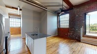 Located in Providence, RI, this industrial-style loft seamlessly blends historic character with modern updates. Original hardwood floors stretch throughout the single-floor layout, reflecting natural light that pours in through large, multi-pane factory-style windows set within striking exposed brick walls. The soaring ceilings are accented by original wood beams and exposed ductwork, reinforcing the space's industrial aesthetic.

The open-concept kitchen is equipped with stainless steel appliances, including a refrigerator, range, and microwave, complemented by white shaker cabinetry and a center island featuring a dark stone countertop and built-in sink. The living area extends naturally from the kitchen, anchored by a ceiling fan and direct access to a private outdoor balcony through a glazed door.

A hallway lined with exposed brick leads to the bedrooms, each fitted with wall-mounted mini-split HVAC units for individual climate control. The primary bathroom offers a dual-sink vanity with dark stone countertops, white cabinetry, round mirrors, and stylish matte black fixtures. Throughout the space, the interplay of raw brick, warm wood, and clean gray walls creates a sophisticated, urban atmosphere distinctive of a thoughtfully converted historic building.