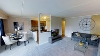 This single-floor space showcases an open-concept design that seamlessly integrates the living, dining, and kitchen areas into one cohesive environment. The living area is finished with plush, light gray carpeting that adds warmth and comfort underfoot, while neutral gray walls create a clean, understated backdrop throughout. A half-wall with a dark countertop serves as a natural divider between the living space and the kitchen, maintaining visual openness while defining each zone.

The kitchen features warm-toned wooden cabinetry, a dark tile backsplash, and black appliances, including a built-in microwave and a range, lending the space a classic, functional aesthetic. A peninsula with seating extends from the kitchen, offering a convenient casual dining option alongside a separate dining area accommodating a small table and chairs.

The living area provides ample room for seating and entertainment, with a media console serving as a focal point. A hallway visible from the main living space suggests additional rooms beyond. Flush-mount ceiling lighting illuminates the interior, and the overall layout is designed to maximize the sense of space across the single level.