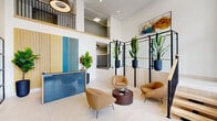 Located at 8680 E Alameda Ave in Denver, Colorado, this professionally designed space makes a striking first impression with its soaring double-height lobby ceiling and expansive floor-to-ceiling glass windows that invite abundant natural light throughout. The welcoming reception area features a bold, color-blocked front desk finished in vibrant blue with warm wood accents, complemented by vertical slatted wall panels in blue and gold tones. A curated seating arrangement of rounded, caramel-upholstered lounge chairs surrounds a fluted wood coffee table, creating an inviting and functional gathering area. Tall tropical plants in dark cylindrical planters are positioned throughout on sleek black metal plant stands, adding a lush, biophilic dimension to the interior. Colorful abstract artwork adorns the bright white walls, contributing to the space's vibrant, creative energy. Glass-walled private offices with black steel-frame partitions line one side of the space, offering transparency while maintaining professional separation. A wood-and-steel staircase with a warm oak handrail connects to an upper-level mezzanine overlooking the lobby below, further enhancing the sense of openness and volume. Light-toned large-format tile flooring runs throughout, tying the design together with a clean, polished finish.