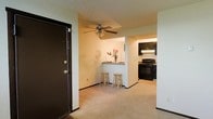 Located at 715 Moon Road in Columbus, Ohio, this single-floor unit offers a practical and comfortable living layout. Upon entry through the dark-finished front door, you are welcomed into a carpeted living area that flows openly into the rest of the space. A ceiling fan overhead provides year-round comfort and adds a classic touch to the main living area. A half-wall breakfast bar with seating for two serves as a natural dividing element between the living space and the kitchen, making it an ideal spot for casual dining or entertaining. The kitchen is equipped with dark cabinetry that contrasts cleanly against lighter walls, and includes black appliances — including a gas range and overhead microwave — lending the space a cohesive, functional aesthetic. Under-cabinet lighting in the kitchen adds warmth and practicality. The neutral color palette throughout, combined with the open sightlines between rooms, creates an inviting and easy-to-personalize atmosphere. The layout is efficiently designed for single-floor living, with each area transitioning naturally into the next.