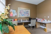 This single-floor space is thoughtfully designed to offer both functionality and a warm, inviting atmosphere. The interior is finished in rich, earthy taupe walls paired with crisp white trim and moldings, creating a cohesive and polished aesthetic throughout.

The dining area is a bright and comfortable spot, anchored by a natural wood table surrounded by upholstered chairs. Large windows fitted with white horizontal blinds allow abundant natural light to filter in, while a glass-paned exterior door provides direct access to the outdoors and extends the sense of openness. A decorative floral arrangement adds a welcoming touch to the space.

Adjacent to the dining area is a built-in workstation that spans one wall, offering ample surface space equipped with a computer, printer, and an array of informational brochures and resource materials. A framed piece of abstract artwork hangs above, adding a refined artistic element. A bulletin board displays community notices and flyers, reinforcing the communal nature of the space.

A small bookcase near the interior glass-paned French door holds a curated selection of reading materials, creating a modest library nook. The French door, featuring multi-pane glass panels and a transom window above, connects this room to a broader interior space visible beyond, hinting at a larger surrounding environment. Neutral carpeting grounds the space and contributes to its warm, approachable character.