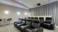 This single-floor screening room is designed to deliver a true cinematic experience within a private setting. The space is outfitted with multiple rows of plush, black leather power recliner seats arranged in a tiered layout, ensuring unobstructed sightlines from every position. Each recliner features integrated cup holders and generous cushioning, prioritizing comfort during extended viewing sessions. A ceiling-mounted projector is centrally positioned to deliver a large, high-quality projection onto the screened wall, which is framed by elegant floor-to-ceiling gray drape curtains that enhance both acoustics and visual presentation. The walls feature a clean, light-toned paneled finish, contributing to the room's polished and refined aesthetic. Recessed ceiling lighting provides subtle ambient illumination, while the neutral flooring ties the interior together cohesively. A small side seating area with casual chairs is visible along one wall, offering additional flexible seating. The overall atmosphere is intimate and purpose-built, combining functional design with upscale finishes to create an exceptional private theater environment.
